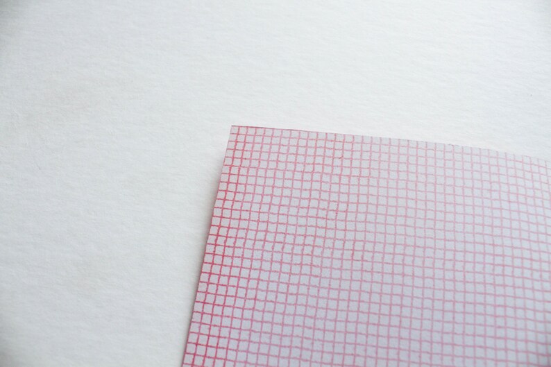 May include: A corner of a white and pink grid pattern on a white background. The grid is composed of thin, red lines forming small squares. The paper has a slightly translucent quality.