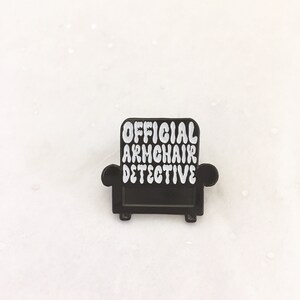 Peut inclure: &Eacute;pinglette en &eacute;mail noir en forme de fauteuil avec le texte "OFFICIAL ARMCHAIR DETECTIVE" en blanc.