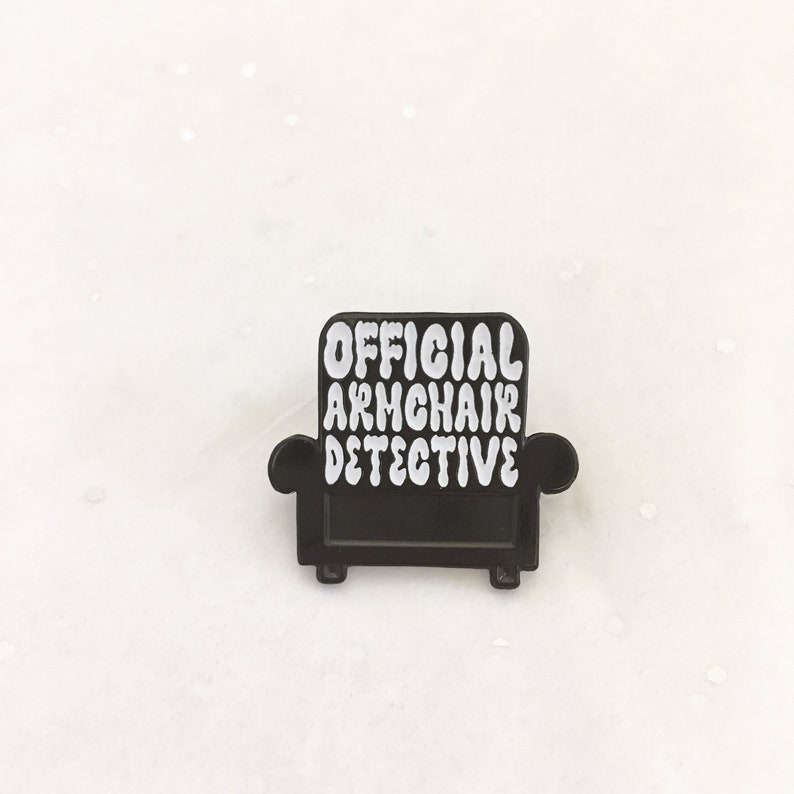 Peut inclure: &Eacute;pinglette en &eacute;mail noir en forme de fauteuil avec le texte "OFFICIAL ARMCHAIR DETECTIVE" en blanc.