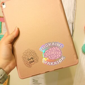 May include: A pink and gold iPad with two stickers on the back. The sticker on the left is a black outline of a person with curly hair and the text "Curly Club". The sticker on the right is a holographic sticker with a pink brain and the text "Migraine Warrior".