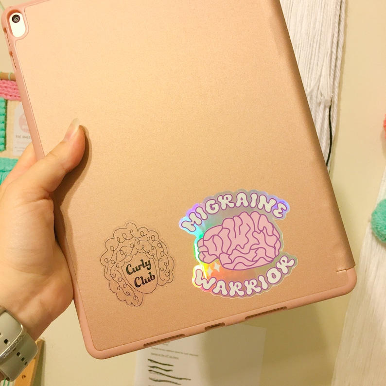 May include: A pink and gold iPad with two stickers on the back. The sticker on the left is a black outline of a person with curly hair and the text "Curly Club". The sticker on the right is a holographic sticker with a pink brain and the text "Migraine Warrior".
