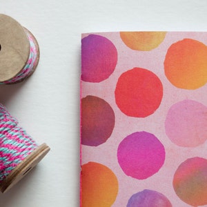 May include: Two wooden spools of twine, one with pink and teal thread, and a notebook with a pink background and large, colorful polka dots. The dots are in shades of orange, red, and purple.