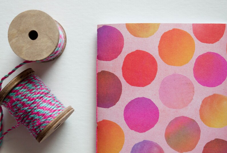 May include: Two wooden spools of twine, one with pink and teal thread, and a notebook with a pink background and large, colorful polka dots. The dots are in shades of orange, red, and purple.