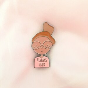 May include: A pink enamel pin featuring a cartoon illustration of a person with brown hair, glasses, and a pink shirt that says "ALWAYS TIRED".