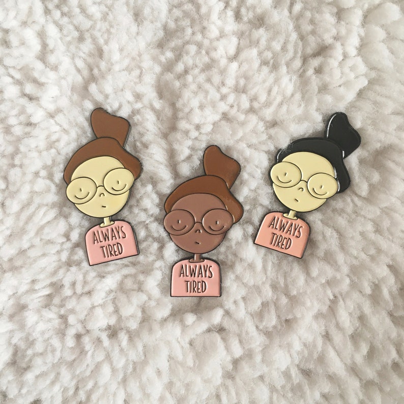 May include: Three enamel pins featuring cartoon illustrations of people with glasses and the text "Always Tired". The pins are on a white fluffy surface.