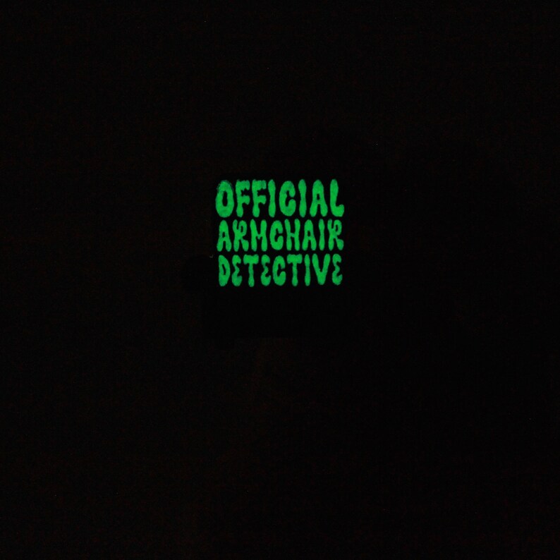 Peut inclure: Une &eacute;pingle verte qui brille dans le noir avec le texte "OFFICIAL ARMCHAIR DETECTIVE" dans une police de caract&egrave;res ludique.