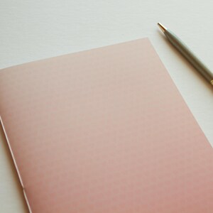 May include: A blank, pink notebook with a subtle gradient effect lies on a white surface. A silver pen with gold accents rests beside the notebook.
