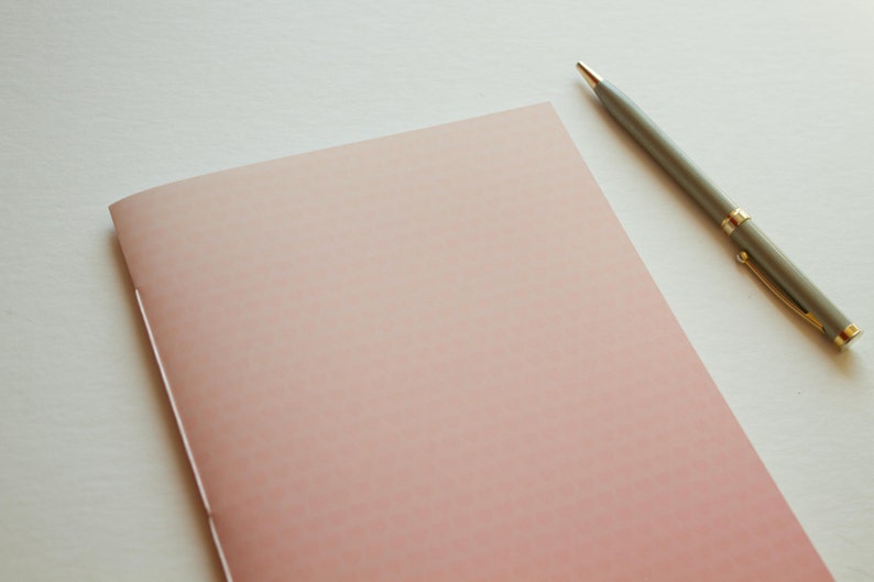 May include: A blank, pink notebook with a subtle gradient effect lies on a white surface. A silver pen with gold accents rests beside the notebook.