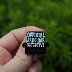 Peut inclure: &Eacute;pinglette en &eacute;mail noir en forme de fauteuil avec le texte "OFFICIAL ARMCHAIR DETECTIVE" en lettres blanches.