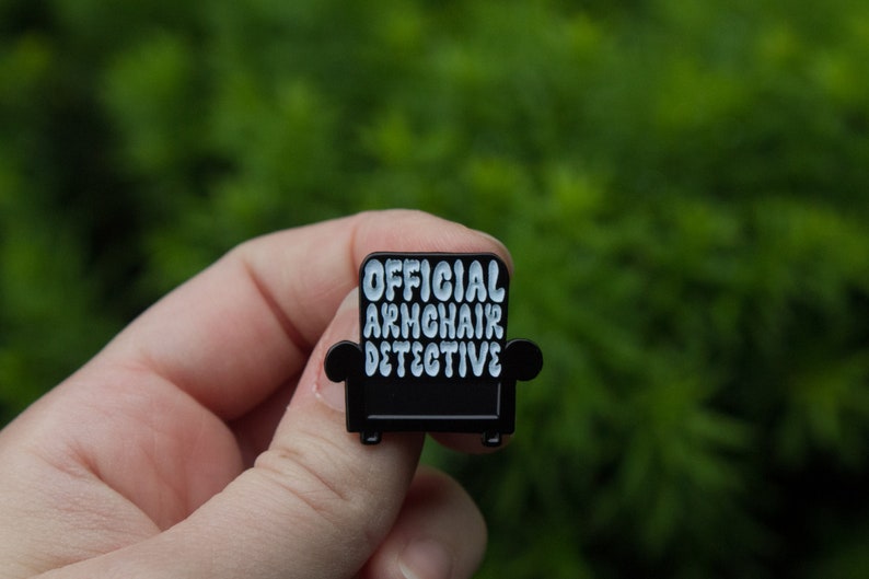 Peut inclure: &Eacute;pinglette en &eacute;mail noir en forme de fauteuil avec le texte "OFFICIAL ARMCHAIR DETECTIVE" en lettres blanches.
