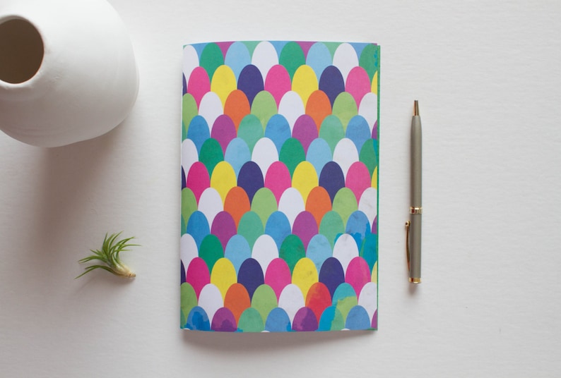 Puede incluir: Un cuaderno colorido con un patr&oacute;n de c&iacute;rculos superpuestos en tonos de rosa, azul, verde, amarillo y naranja. El cuaderno tiene una cubierta turquesa.