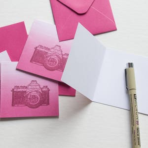 May include: Pink and white blank greeting cards with a vintage camera design. The cards are stacked on top of each other. A brown pen is lying on top of the cards.