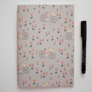May include: A grey notebook with a repeating pattern of bicycles and orange flowers. The notebook is closed and has a black pen next to it.