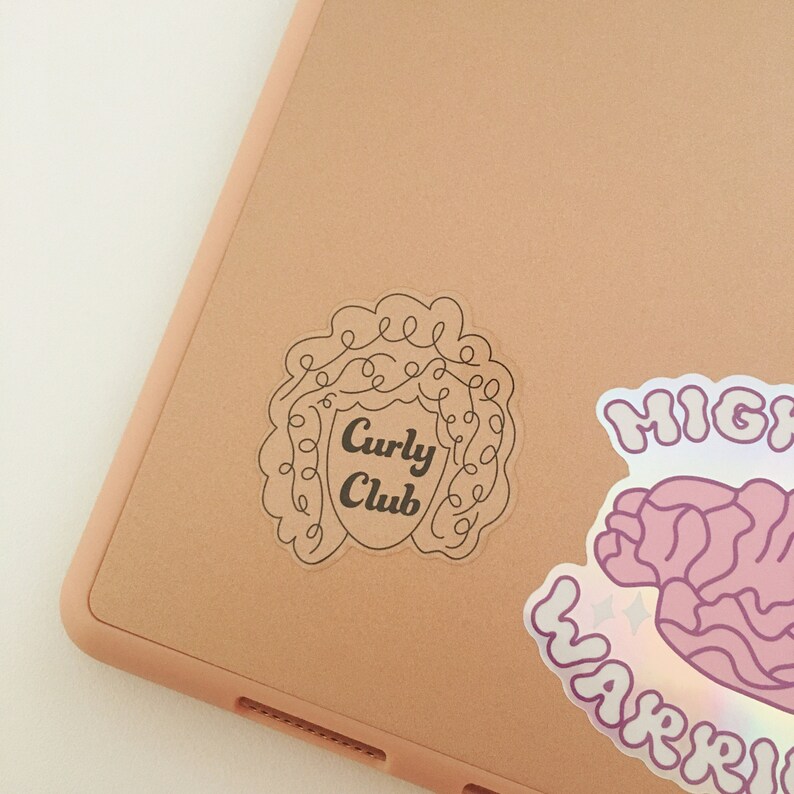 May include: A brown laptop with a sticker of a curly haired person with the text "Curly Club" on it. There is another sticker on the laptop that says "Mighty Warrior" with a pink and purple design.