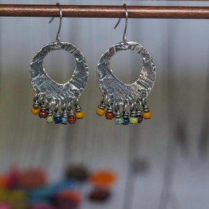 Puede incluir: Pendientes de aro de plata con textura martillada y una franja colgante de cuentas de colores. Las cuentas son amarillas, naranjas, rojas, azules y verdes.