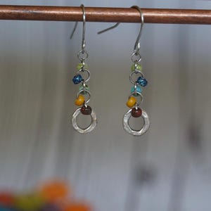 Puede incluir: Pendientes de plata con un dije circular y cuentas de colores en tonos azul, verde, amarillo y marrón.