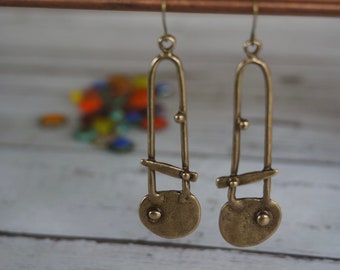 Könnte beinhalten: Ein Paar lange, goldfarbene Ohrringe mit geometrischem Design. Jeder Ohrring hat eine runde Scheibe mit einem kleinen, erhabenen Punkt in der Mitte.