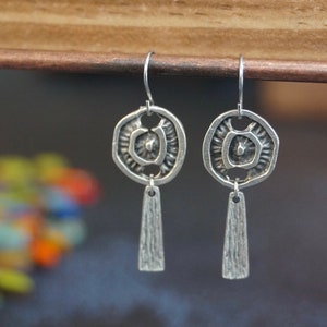 Puede incluir: Pendientes de plata con un diseño circular texturizado y un largo y delgado colgante texturizado. Los pendientes están colgados de una superficie de madera.