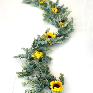 May include: A garland of artificial greenery with small round leaves and yellow sunflower blossoms. The garland is arranged in a winding pattern.