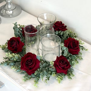 May include: A centerpiece featuring clear glass vases surrounded by a ring of faux greenery and deep red roses. The arrangement sits on a white surface, creating an elegant display for events or home decor.