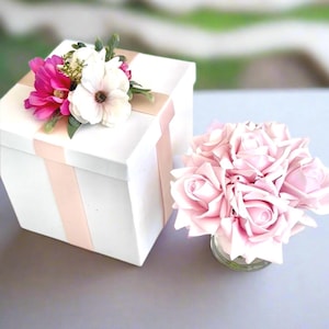 Puede incluir: Una caja de regalo blanca con una cinta rosa y un arreglo floral. Las flores son rosas y blancas con hojas verdes. Un pequeño ramo de rosas rosas está en un jarrón de cristal.