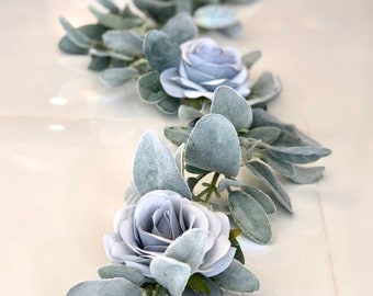 Puede incluir: Una guirnalda de rosas artificiales de color azul polvoriento y hojas verde grisáceas. La guirnalda es perfecta para añadir un toque de elegancia a cualquier espacio.
