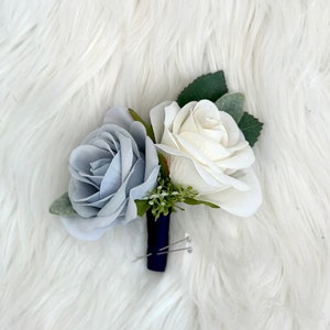May include: A blue and white artificial flower boutonniere with a navy blue pin. The boutonniere is made of two roses, one blue and one white, with green leaves and small white flowers.