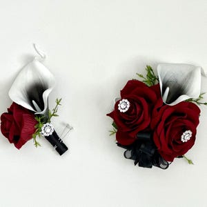 May include: A set of two boutonnieres featuring a single white calla lily and red roses with black ribbon and rhinestone accents.