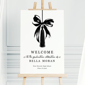 May include: A white sign on a wooden easel with black text and a black bow and tassel illustration. The text reads "WELCOME to the graduation celebration of BELLA MORAN West Waverly High School Class of 2026."