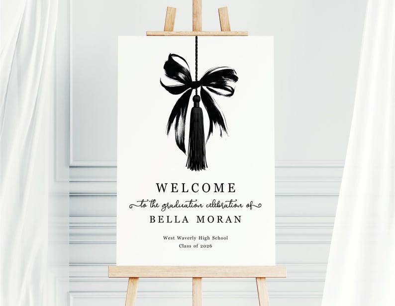 May include: A white sign on a wooden easel with black text and a black bow and tassel illustration. The text reads "WELCOME to the graduation celebration of BELLA MORAN West Waverly High School Class of 2026."