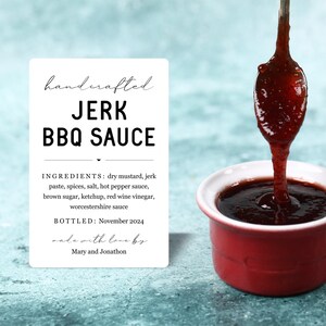 K&ouml;nnte beinhalten: Ein wei&szlig;es Etikett mit schwarzem Text, auf dem "handcrafted JERK BBQ SAUCE" steht. Das Etikett listet die Zutaten auf und gibt an, dass die Sauce im November 2024 abgef&uuml;llt wurde. Das Etikett enth&auml;lt auch den Text "made with love by Mary and Jonathon". Ein L&ouml;ffel mit roter Sauce tropft in eine rote Sch&uuml;ssel.