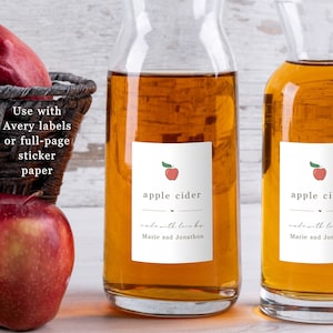 Könnte beinhalten: Zwei Glasflaschen gefüllt mit Apfelsaft, jede mit einem weißen Etikett, auf dem "Apfelsaft" und "mit Liebe gemacht von Marie und Jonathon" steht. Die Etiketten zeigen einen roten Apfel mit einem grünen Blatt.