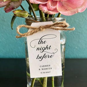 May include: A clear glass vase filled with pink flowers and green stems, featuring a white rectangular paper tag tied with brown twine. The tag displays the black printed text: the night before, CARMEN & MARCUS, 6.19.2027, in a mix of script and serif fonts.