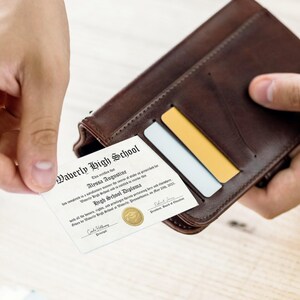 May include: A white diploma with a gold seal, reading "Waverly High School" and "Alyssa Augustine High School Diploma" with the date "May 2021". The diploma is being pulled out of a brown leather wallet.
