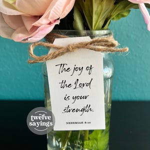 May include: A clear glass Mason jar vase filled with pink and cream-colored flowers and greenery. A white tag tied with twine reads "The joy of the Lord is your strength." The text is from Nehemiah 8:10. The vase has a black surface.