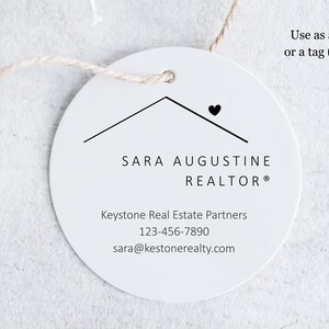 Pu&ograve; includere: Un'etichetta rotonda bianca con una grafica a forma di tetto di casa e un cuore. L'etichetta include il testo "SARA AUGUSTINE REALTOR&reg;", le informazioni di contatto e la frase "Use as a sticker or a tag (or both)." &Egrave; attaccato un cordino.