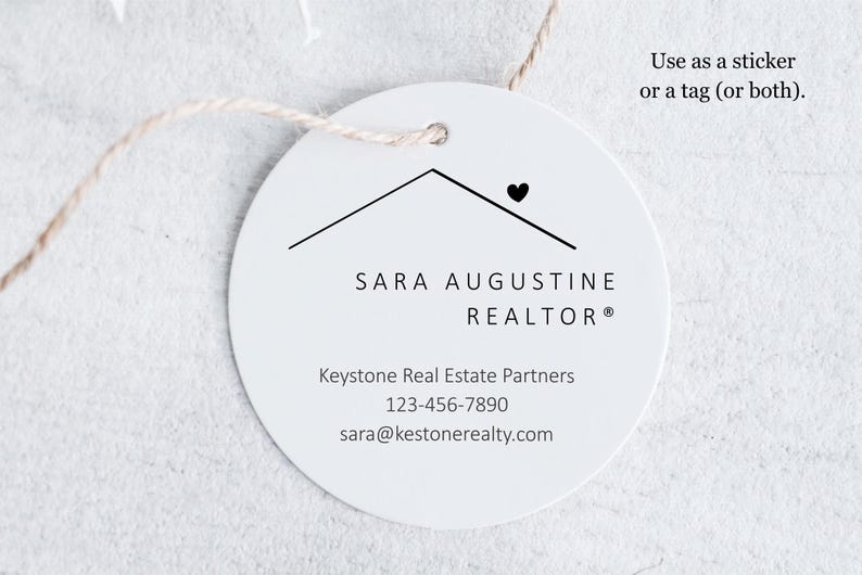 Pu&ograve; includere: Un'etichetta rotonda bianca con una grafica a forma di tetto di casa e un cuore. L'etichetta include il testo "SARA AUGUSTINE REALTOR&reg;", le informazioni di contatto e la frase "Use as a sticker or a tag (or both)." &Egrave; attaccato un cordino.