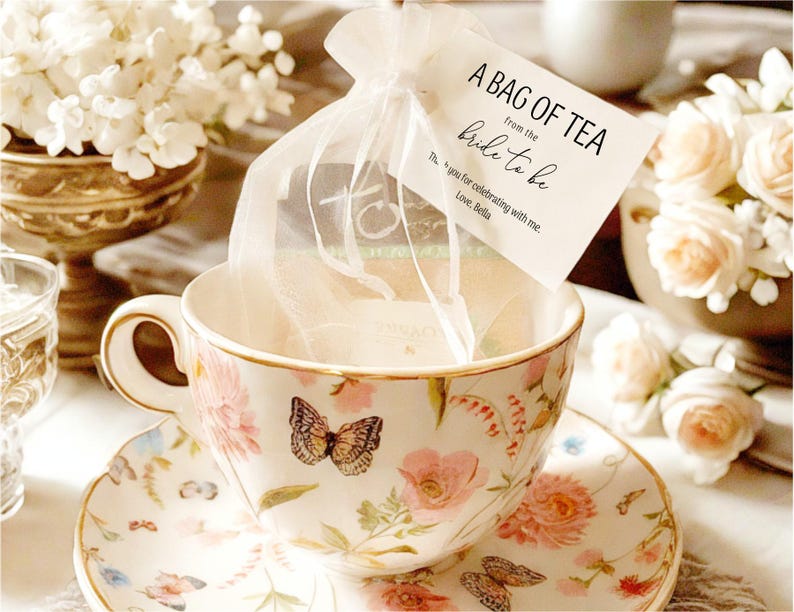 Puede incluir: Una taza de t&eacute; y un platillo blancos con un patr&oacute;n floral, un dise&ntilde;o de mariposa y un borde dorado. Una peque&ntilde;a bolsa blanca con una etiqueta que dice "A Bag of Tea from the Bride to Be" est&aacute; sentada en la taza de t&eacute;.