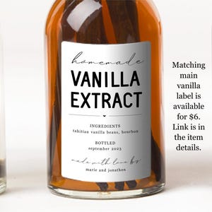 May include: A clear glass bottle filled with amber liquid and dark vanilla beans, featuring a white rectangular label with black text that reads Homemade Vanilla Extract, Ingredients Tahitian vanilla beans, bourbon, Bottled September 2023, made with love by marie and jonathon.