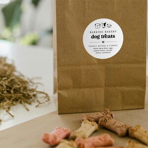 May include: A brown paper bag with a white label that reads "Barking Bakery Dog Treats." The label lists ingredients: peanut butter, carrot, whole wheat flour, eggs, peanut butter, honey, and cornstarch. Several dog treats are scattered in front of the bag.