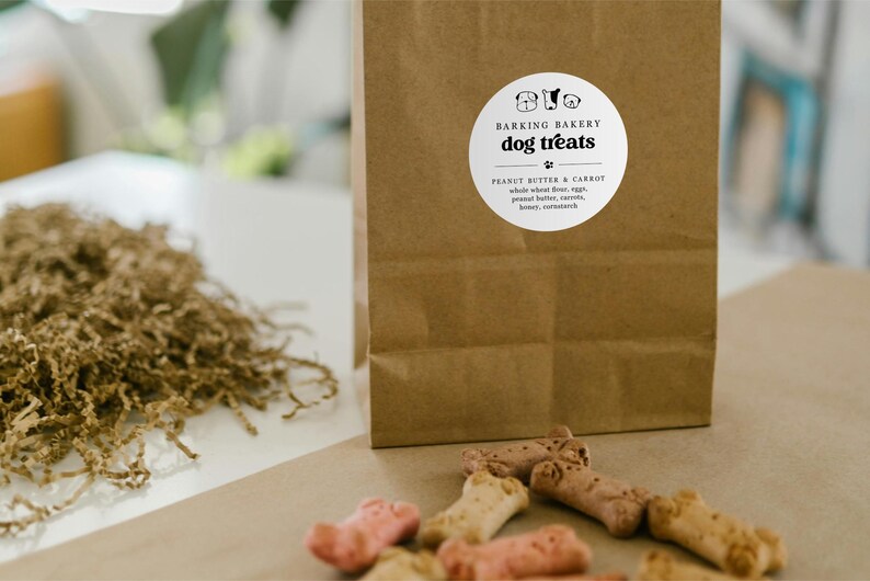 May include: A brown paper bag with a white label that reads "Barking Bakery Dog Treats." The label lists ingredients: peanut butter, carrot, whole wheat flour, eggs, peanut butter, honey, and cornstarch. Several dog treats are scattered in front of the bag.