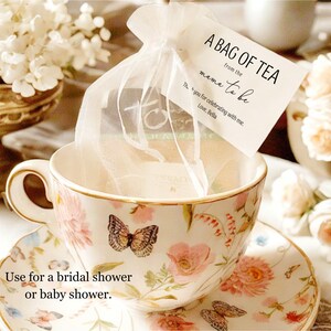 Puede incluir: Una bolsita de t&eacute; blanca en una bolsa blanca transparente con una taza de t&eacute; y un platillo blancos con dise&ntilde;os florales. La bolsita de t&eacute; tiene una etiqueta que dice "A Bag of Tea from the Mom-to-be. Thanks for celebrating with me. Love, Bella."  Util&iacute;zala para una despedida de soltera o un baby shower.