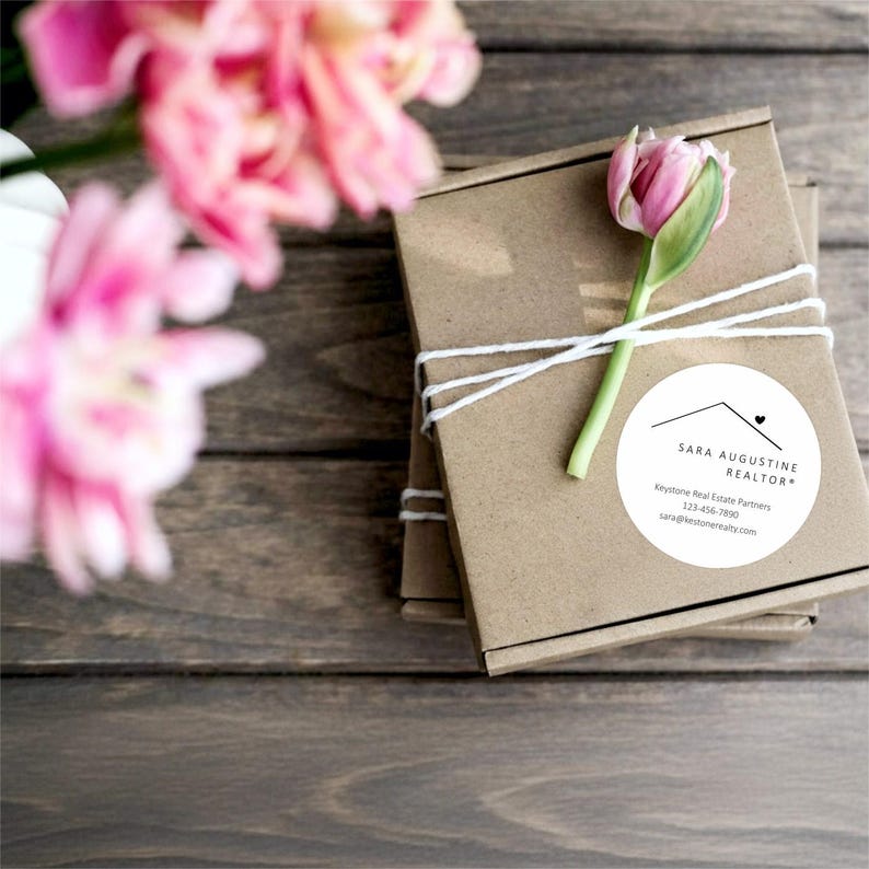 Pu&ograve; includere: Due piccole scatole di cartone marrone legate con spago bianco, sormontate da un tulipano rosa. Un adesivo circolare bianco sulla scatola recita "SARA AUGUSTINE REALTOR" con informazioni di contatto. Fiori rosa sullo sfondo.