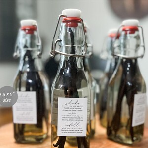 May include: Clear glass swing-top bottles filled with liquid and dark vanilla beans. Each bottle features a white rectangular label with the text: Shake every now and again, use the little black specs, refill with vodka once low. Label size is 3.8 x 5.1 cm.