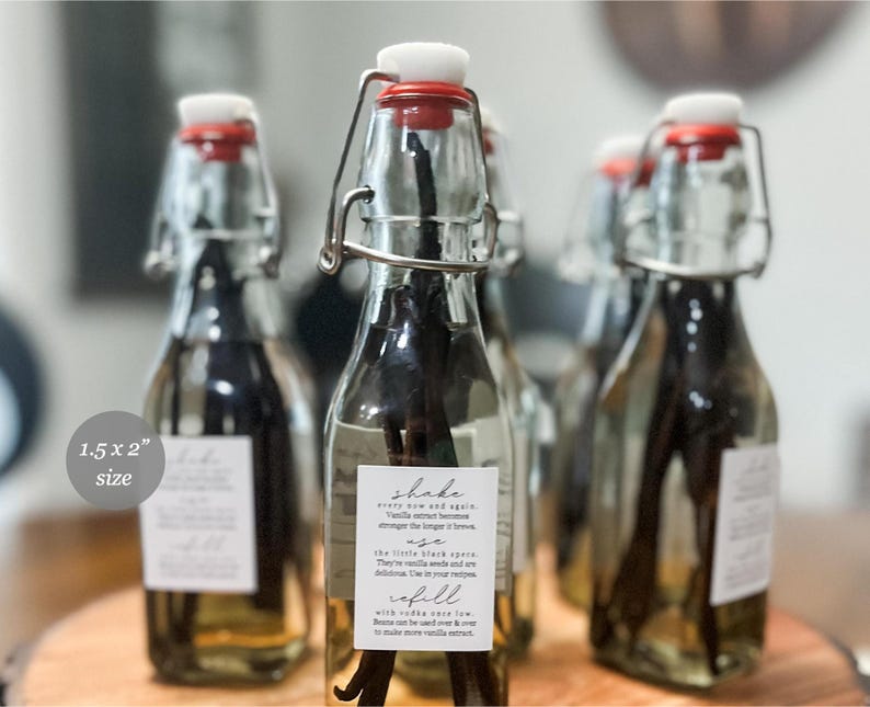 May include: Clear glass swing-top bottles filled with liquid and dark vanilla beans. Each bottle features a white rectangular label with the text: Shake every now and again, use the little black specs, refill with vodka once low. Label size is 3.8 x 5.1 cm.
