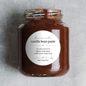 May include: A glass jar filled with homemade vanilla bean paste. The jar has a white round label with the text "homemade vanilla bean paste" and a list of ingredients: "tahitian vanilla beans, vanilla extract, maple syrup". The label also includes the text "made with love by Page Augustine".
