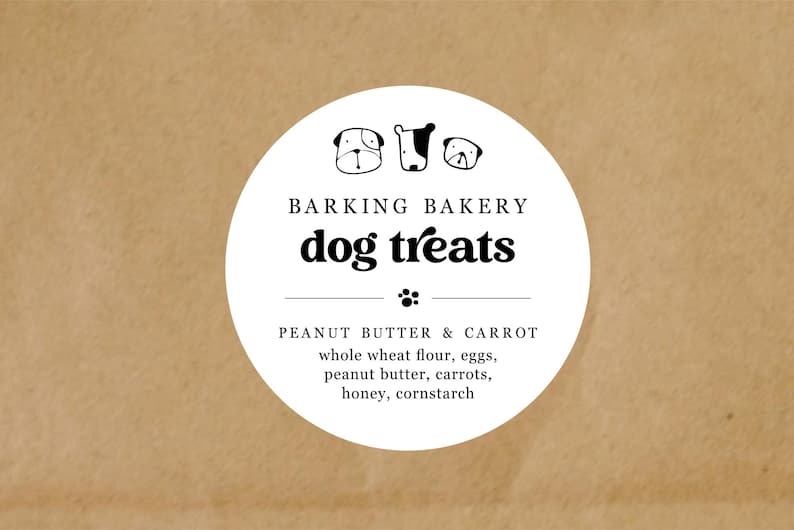 May include: A white circular label on a brown background. The label reads "BARKING BAKERY dog treats" with dog illustrations. Below, it lists ingredients: peanut butter & carrot, whole wheat flour, eggs, peanut butter, carrots, honey, and cornstarch.