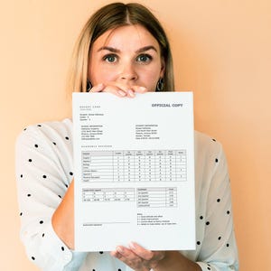 May include: A woman holds up a report card with the words "OFFICIAL COPY" at the top. The report card shows the student's name, grade, school information, and academic record.