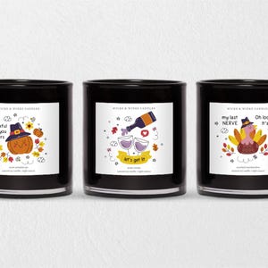 May include: Three black glass candles with white labels featuring humorous illustrations. One candle has a pumpkin wearing a witch hat, another has wine glasses and a bottle, and the third has a turkey. Text includes "thankful for you fuckers", "let's get it", and "Oh look... It's on fire".