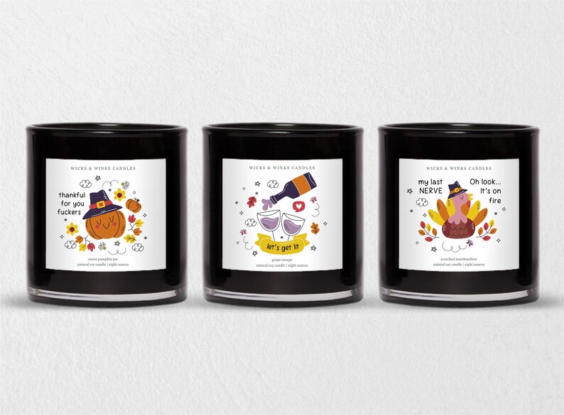 May include: Three black glass candles with white labels featuring humorous illustrations. One candle has a pumpkin wearing a witch hat, another has wine glasses and a bottle, and the third has a turkey. Text includes "thankful for you fuckers", "let's get it", and "Oh look... It's on fire".